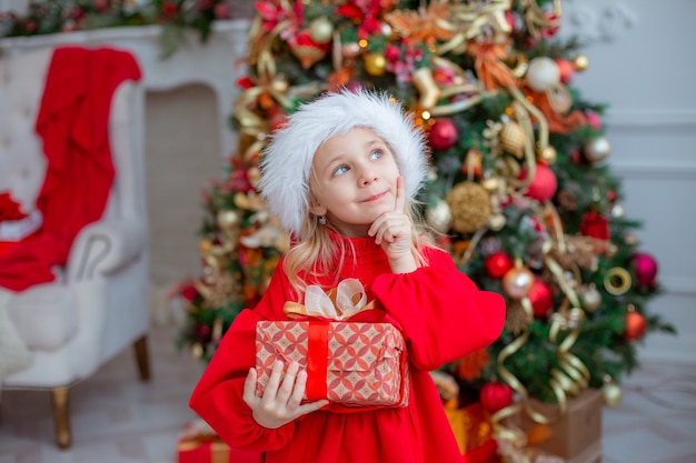 Une petite fille dans un bonnet de Noel tient un cadeau près de l'arbre de Noël à la maison