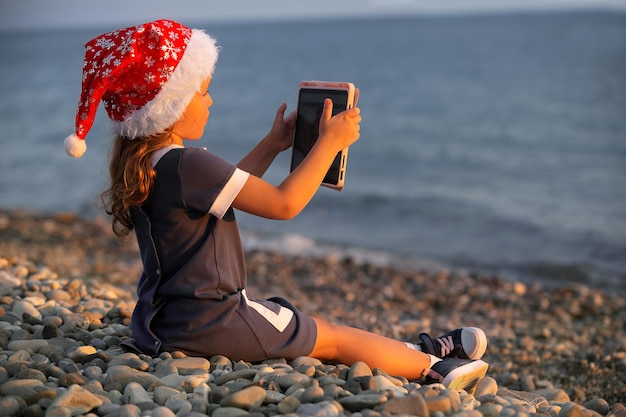une petite fille dans un bonnet rouge est assise au bord de la mer au coucher du soleil et regarde des dessins animés sur une tablette