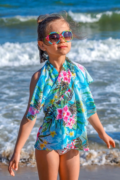 Petite fille dans une cape de plage bleue et lunettes de soleil sur fond de vagues de la mer