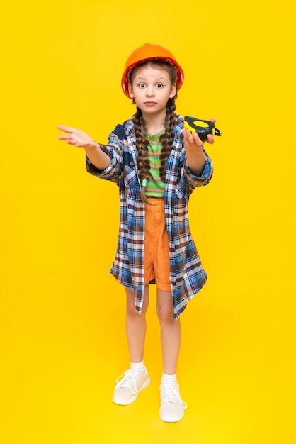 Une petite fille dans un casque de construction orange tient un ruban à mesurer Le concept de rénovation dans la chambre des enfants L'enfant est un ingénieur Vertical Pleine hauteur Fond isolé jaune