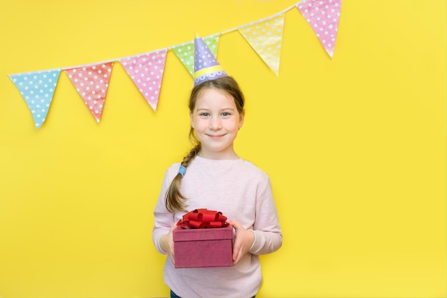 Petite fille dans une casquette tient un cadeau avec un arc dans ses mains sur un fond jaune décoré de drapeaux festifs le concept de cadeaux de vacances