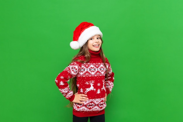 Une petite fille dans un chandail rouge avec des rennes et un chapeau de Père Noël attendant les vacances sur un fond vert isolé Le concept de Noël