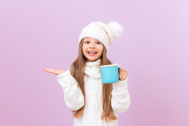 Une petite fille dans un chapeau d'hiver tient une tasse avec un verre et l'autre pointe vers le côté.