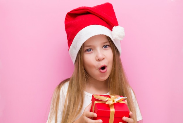 petite fille dans un chapeau de Noël rouge présente une boîte rouge avec un arc en or, un cadeau. Des sourires. La nouvelle année est 2021. Gros plan