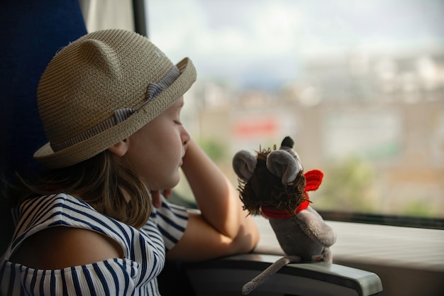 une petite fille dans un chapeau de paille avec un jouet est assise près de la fenêtre dans le train