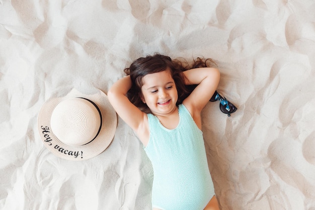 Une petite fille dans un chapeau de paille et des lunettes de soleil prend un bain de soleil sur une plage de sable
