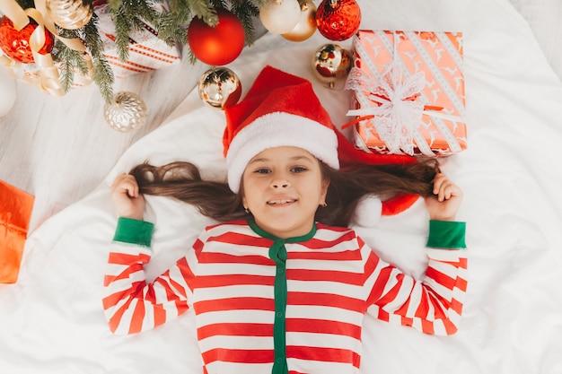 Petite fille dans un chapeau de père Noël se trouve près de l'arbre de Noël à la maison. vue d'en-haut. Noël.