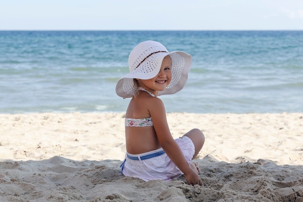 Petite fille dans un chapeau de plage blanc et bikini est assise sur le sable au bord de la mer