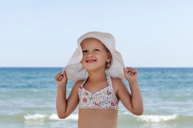 Petite fille dans un chapeau de plage blanc et bikini tenant un chapeau sur un fond de mer et de ciel bleus