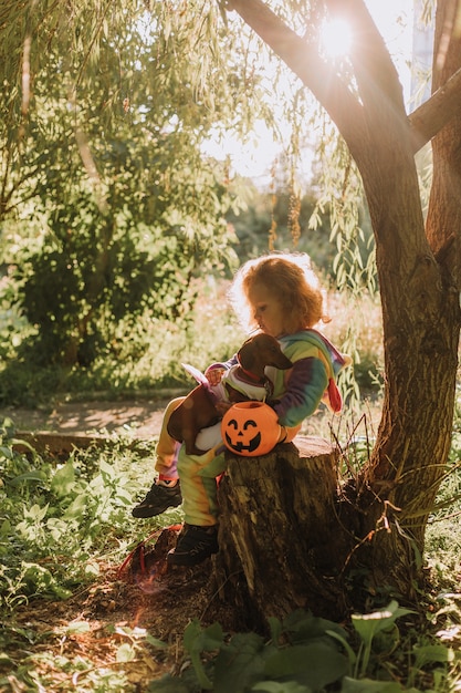 Petite fille dans un costume d'Halloween de licorne arc-en-ciel et un teckel dans une robe avec un panier de citrouille