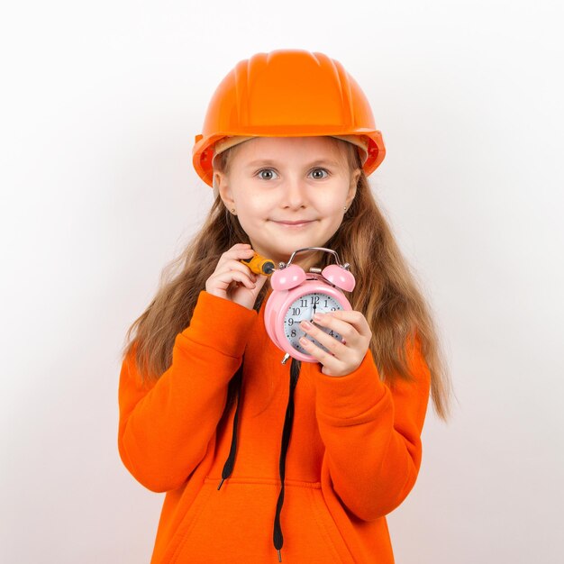 Une petite fille dans un costume orange et un casque de construction orange Fixe le réveil avec un tournevis Fond blanc