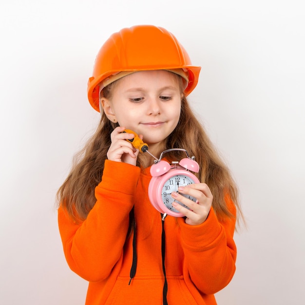 Une petite fille dans un costume orange et un casque de construction orange Fixe le réveil avec un tournevis Fond blanc