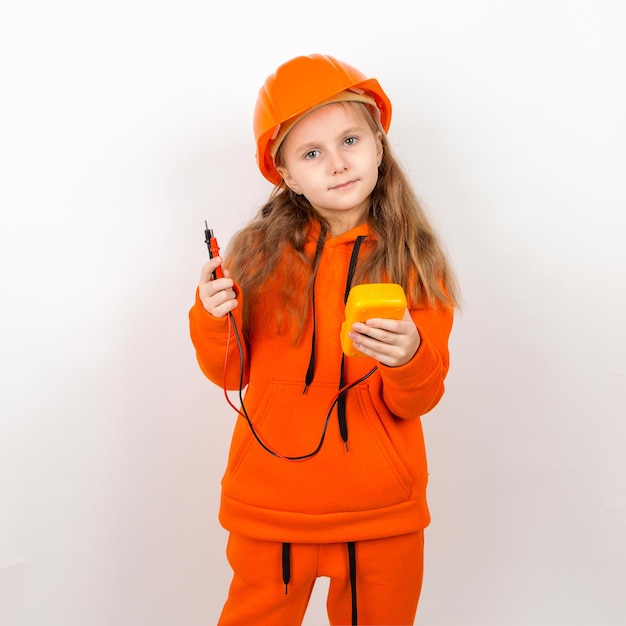 Une petite fille dans un costume orange et un casque de construction tenant un multimètre concept d'un jeune électricien Portrait fond blanc Fête du travail