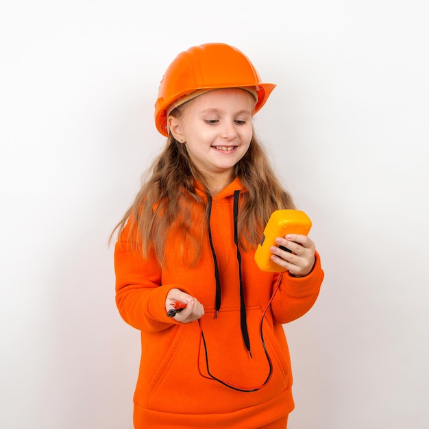 Une petite fille dans un costume orange et un casque de construction tenant un multimètre concept d'un jeune électricien Portrait fond blanc Fête du travail