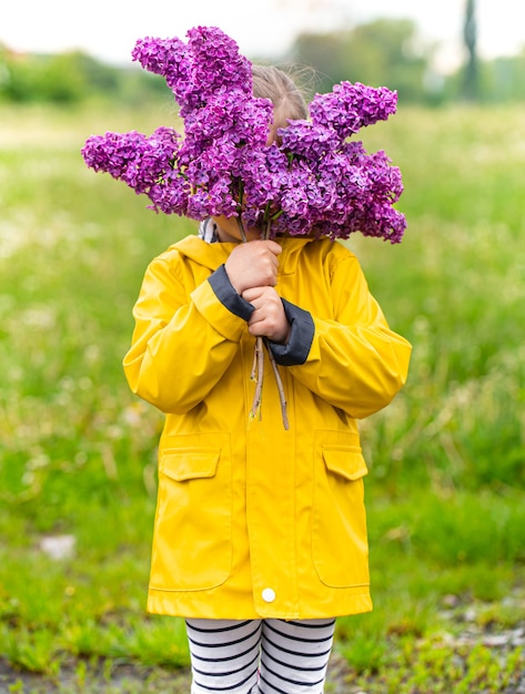 Petite fille dans un imperméable jaune tient un bouquet de fleurs lilas sur l'herbe se bouchent.