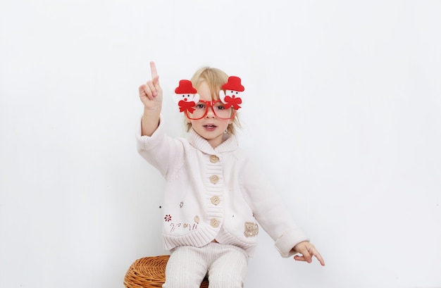 Une petite fille dans des lunettes de Noël drôles avec des bonhommes de neige est assise contre un mur blanc.