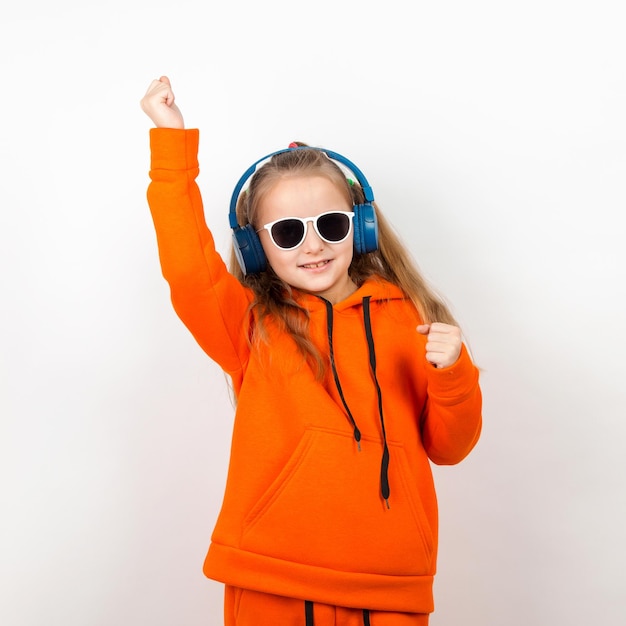 Une petite fille dans des lunettes de soleil à capuche orange et un casque bleu écoute de la musique Portrait sur fond blanc