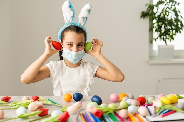 Une petite fille dans un masque médical peint des oeufs de Pâques avec un motif. Rester à la maison.