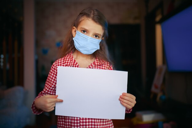 Petite fille dans un masque de protection, le pyjama tient une feuille de papier vierge d'une main et l'autre pointe vers la feuille en se tenant debout dans la pièce à la maison, Protection contre le coronavirus