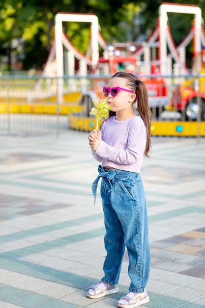 Une petite fille dans un parc d'attractions en été souffle sur une toupie près des carrousels en lunettes de soleil, le concept des vacances d'été et des vacances scolaires