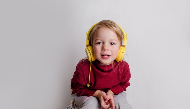 Une petite fille dans un pull rouge écoute de la musique avec des écouteurs et rit sur fond blanc.