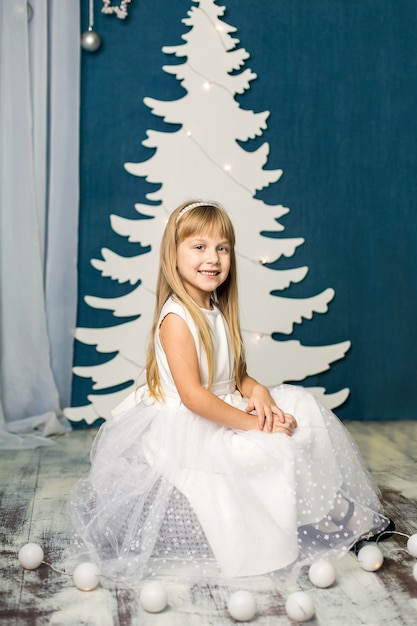 Petite Fille Dans Une Robe Blanche Est Assise Sur Une Bouffée D'un Décor D'arbre De Noël Blanc Artificiel