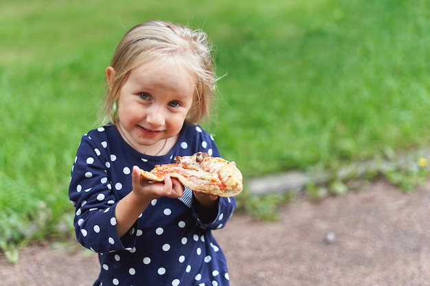 Une petite fille dans une robe bleue à pois blancs présente une grande part de pizza à la caméra