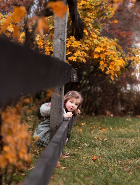 Une petite fille dans une robe à carreaux donne sur une clôture en bois dans un parc en automne.