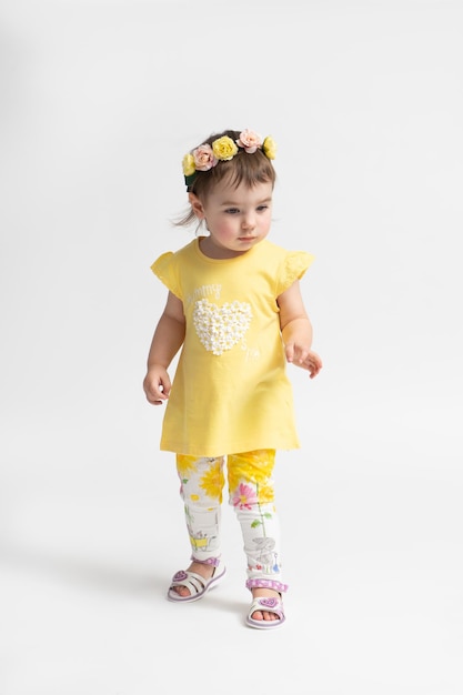 Petite fille dans une robe jaune décontractée pour enfants avec une couronne de fleurs sur la tête posant sur un fond blanc