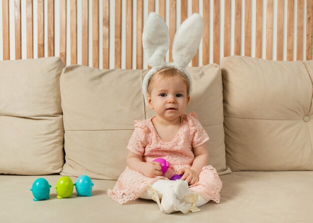 Petite fille dans une robe et des oreilles de lapin est assise sur un canapé avec des œufs colorés