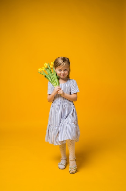 Une petite fille dans une robe rayée se dresse avec un bouquet de tulipes jaunes sur jaune