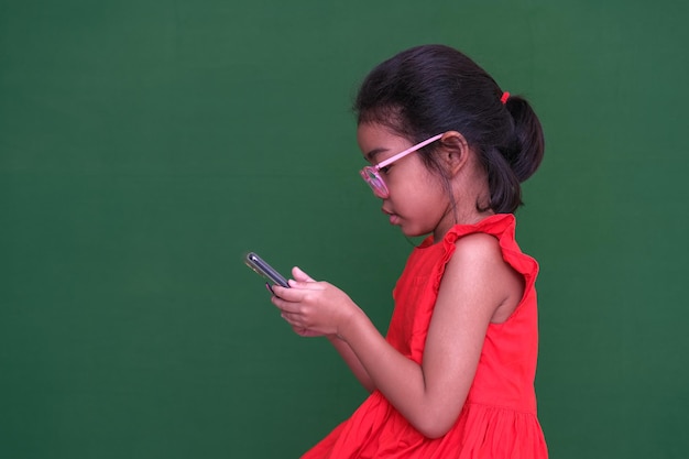 Petite fille dans sa robe rouge et ses lunettes occupées à jouer à un jeu sur son gadget électronique Photo prise sur fond vert foncé