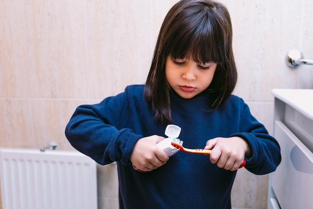 Petite fille dans un sweat-shirt bleu foncé, mettant du dentifrice sur sa brosse à dents dans la salle de bain. Concept de brossage des dents, d'hygiène et d'enfance.