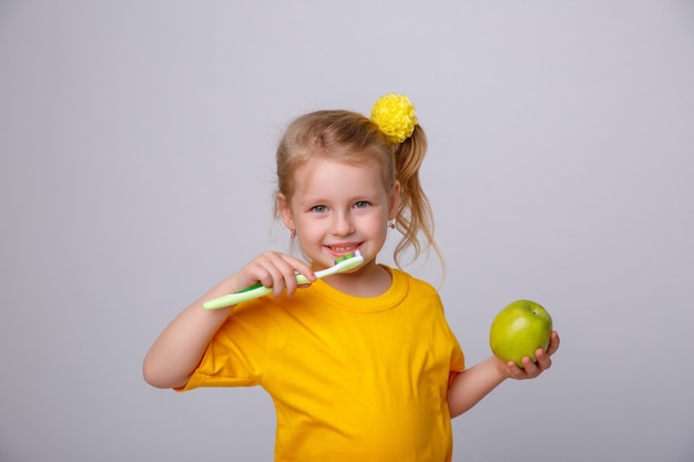 Une petite fille dans un T-shirt jaune tient une brosse à dents et une pomme sur un fond blanc