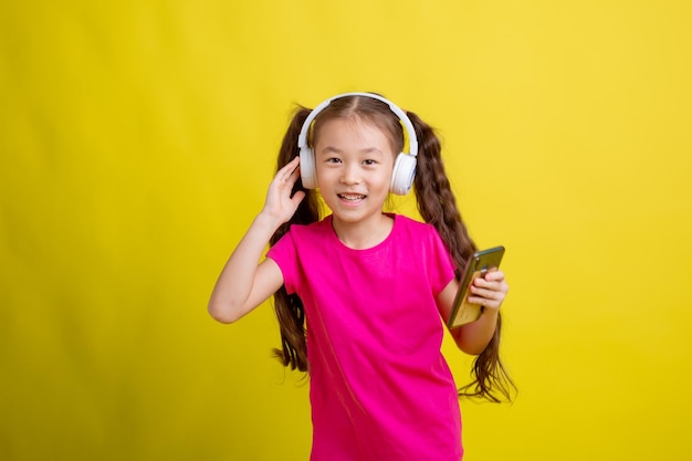 Une petite fille dans un T-shirt rose avec des écouteurs et un téléphone portable sur fond jaune