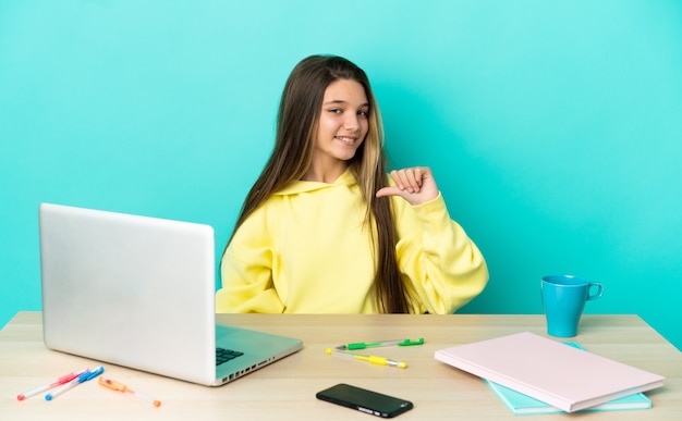 Petite fille dans une table avec un ordinateur portable sur fond bleu isolé fier et satisfait de lui-même