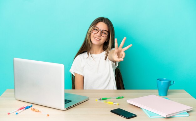 Petite fille dans une table avec un ordinateur portable sur fond bleu isolé heureux et comptant trois avec les doigts