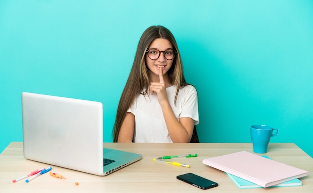 Petite fille dans une table avec un ordinateur portable sur fond bleu isolé montrant un signe de silence geste mettant le doigt dans la bouche