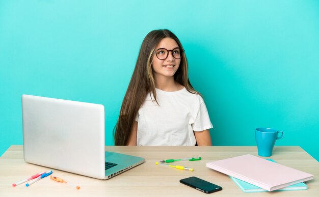 Petite fille dans une table avec un ordinateur portable sur fond bleu isolé en riant