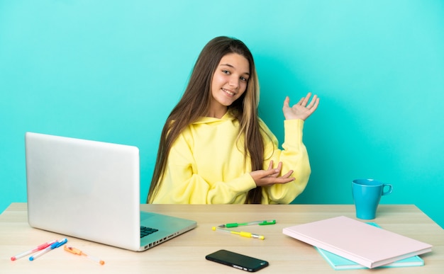 Petite fille dans une table avec un ordinateur portable sur fond bleu isolé tendant les mains sur le côté pour inviter à venir