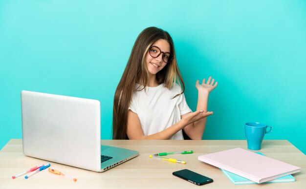 Petite fille dans une table avec un ordinateur portable sur fond bleu isolé tendant les mains sur le côté pour inviter à venir