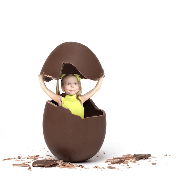 Petite fille dans une veste jaune comme un poulet éclos d'un œuf de Pâques en chocolat sur un fond blanc copie espace collage minimalisme Enfant dans un œuf de Pâques en chocolat