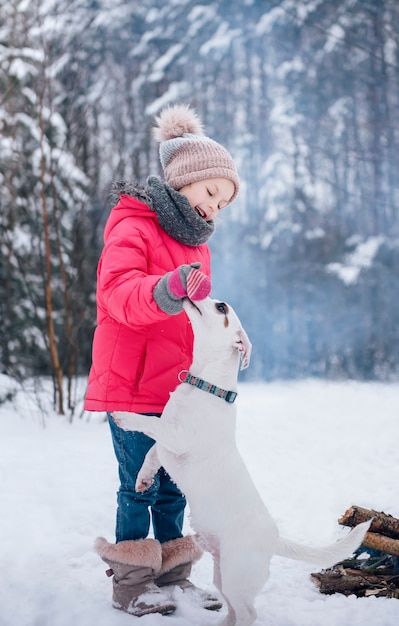 Petite fille dans une veste lumineuse joue dans la forêt enneigée d'hiver avec son chien jack russell terrier