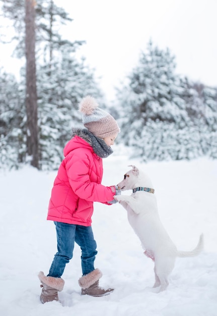 Petite fille dans une veste lumineuse joue dans la forêt enneigée d'hiver avec son chien jack russell terrier