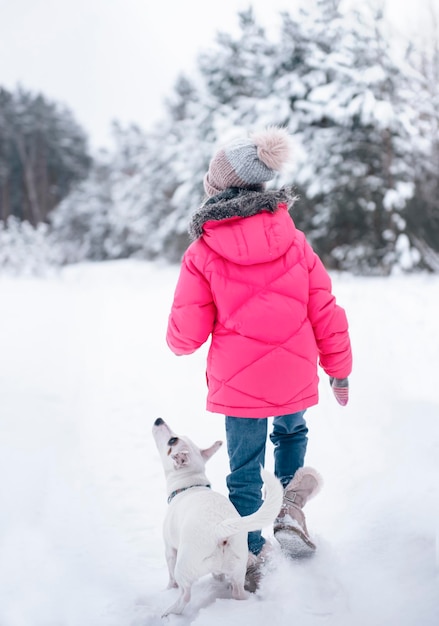 Petite fille dans une veste lumineuse joue dans la forêt enneigée d'hiver avec son chien jack russell terrier