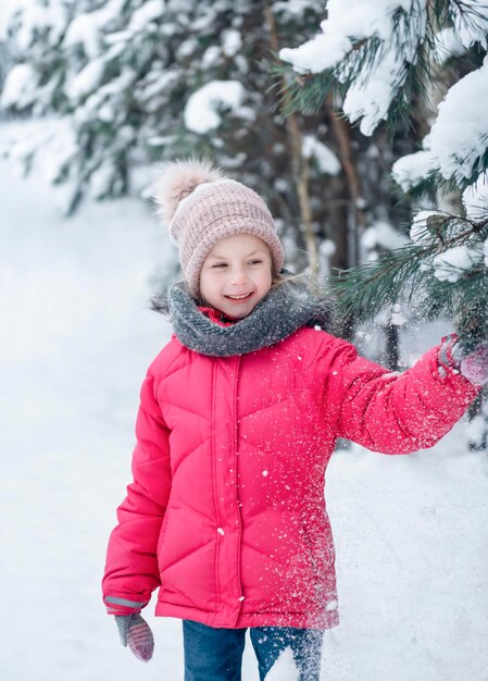 Une petite fille dans une veste lumineuse joue dans la forêt enneigée d'hiver.