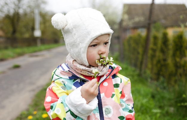 Une petite fille dans une veste lumineuse se tient dehors au printemps et tient une branche avec des fleurs blanches printanières dans ses mains.