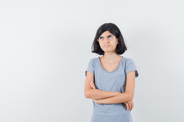 Petite fille debout avec les bras croisés en t-shirt et à la recherche hésitante. vue de face.