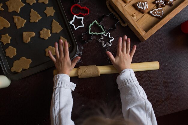 Une petite fille déroule la pâte avec un rouleau à pâtisserie sur la table et prépare des biscuits de Noël