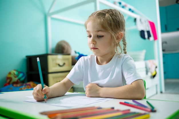 Petite fille dessine assise à une table dans une pièce sur fond de mur